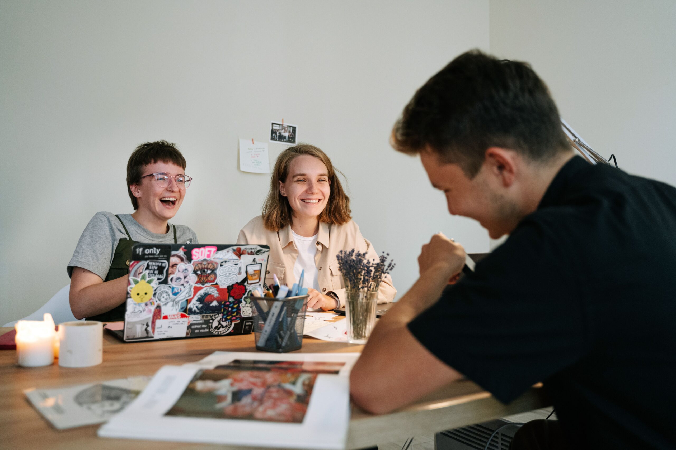 WebAndThat team laughing candidly around desks with mismatched tea mugs, stickered laptops, and a tiny knitted hat on a desk plant